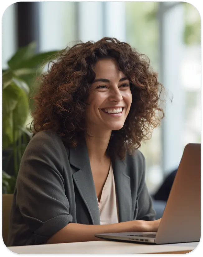 A smiling woman with curly hair working on her laptop in a bright office setting.